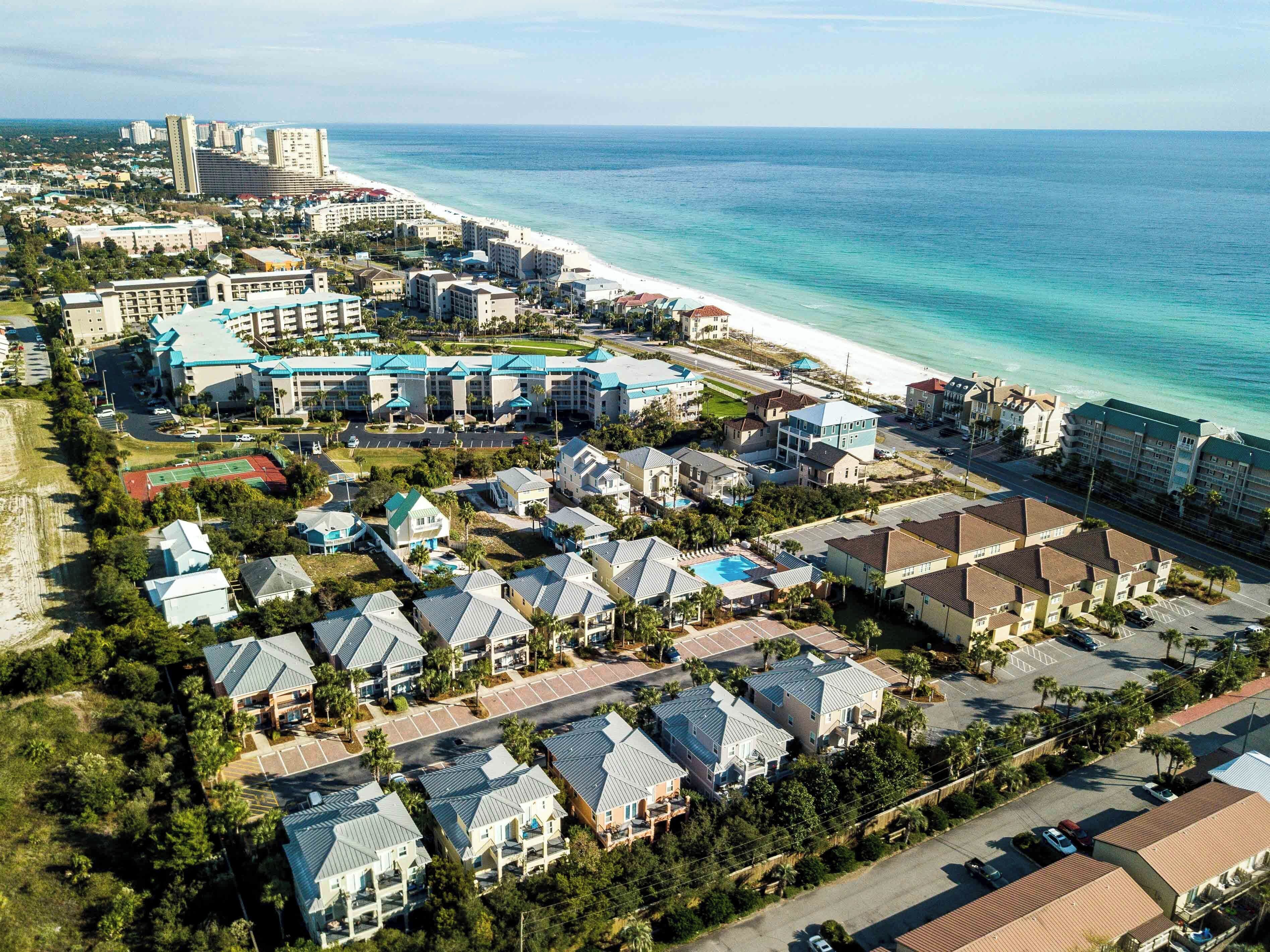 An aerial view of a coastal resort area with multiple buildings, including a large hotel with a blue roof and a smaller residential neighborhood with single-family homes.