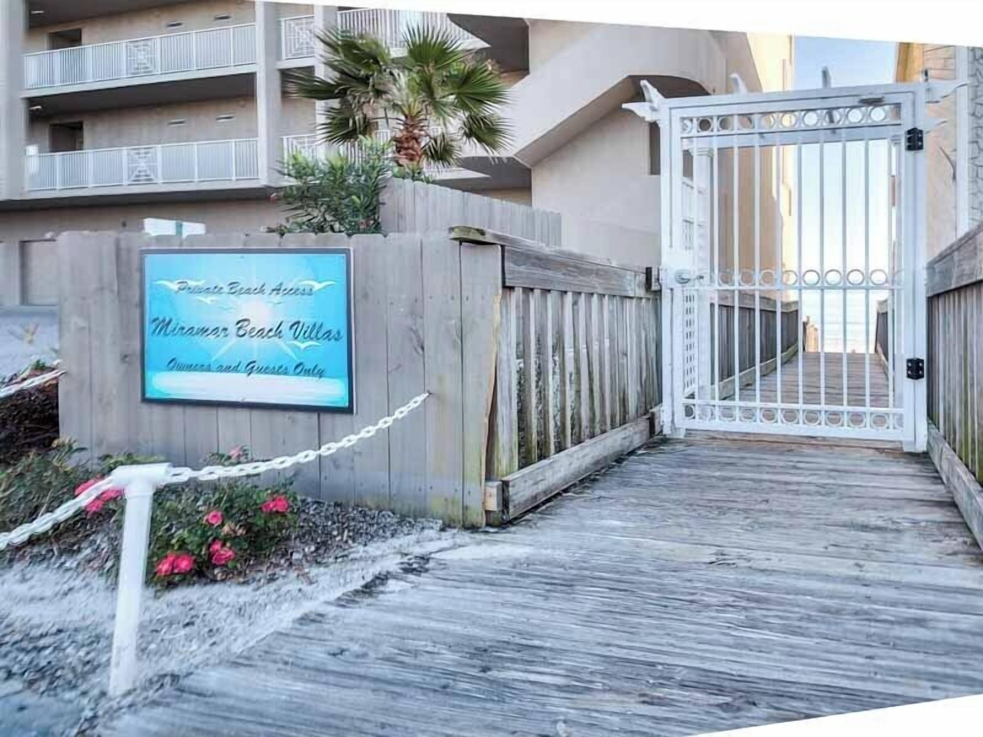 Een hek open naar een privéstrandtoegang met een bord waarop 'Miramar Beach Villas' en 'Alleen voor eigenaren en gasten' staat.
