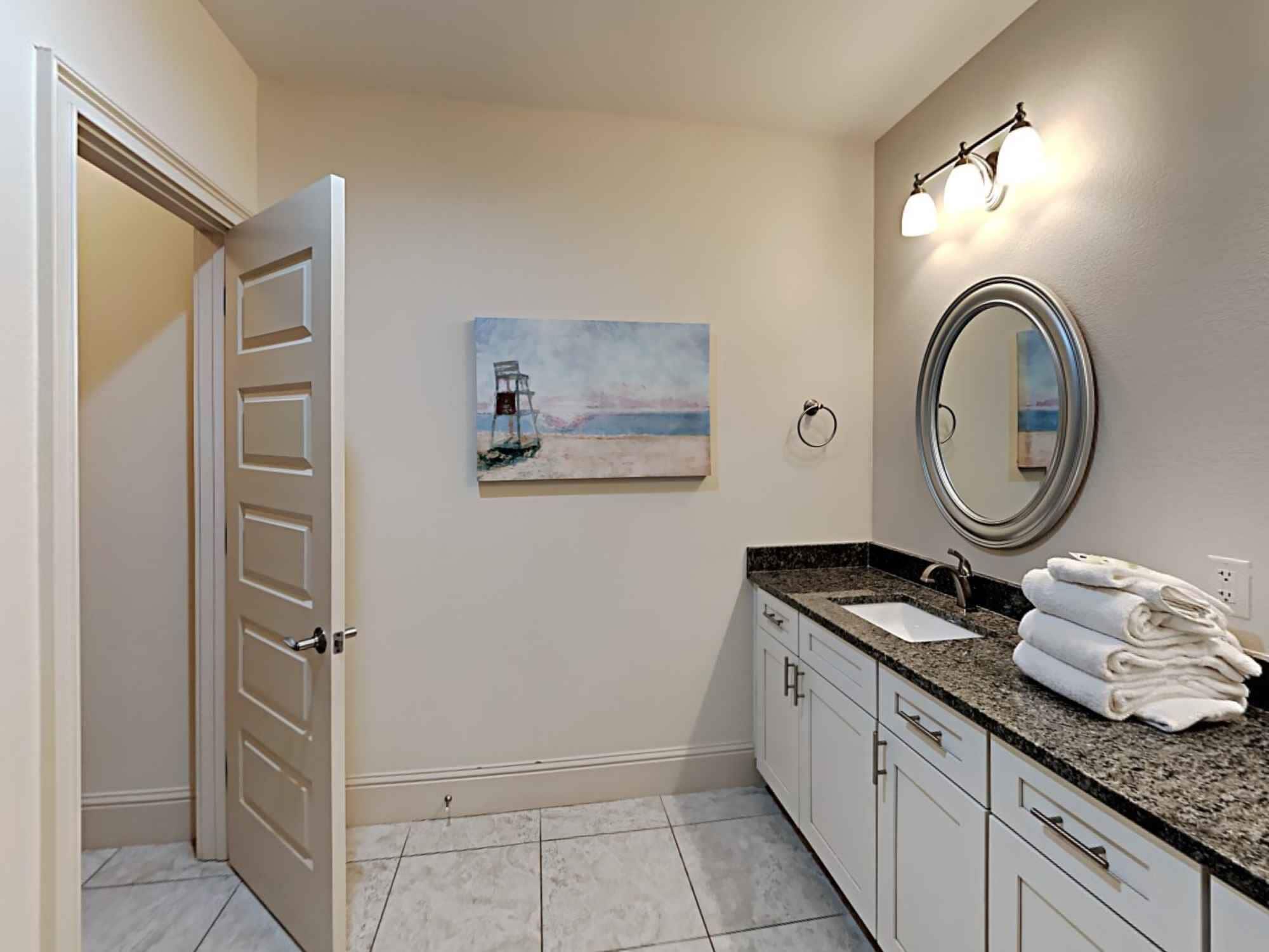 A bathroom with a painting of a beach scene, a round mirror, and a stack of towels on the counter.
