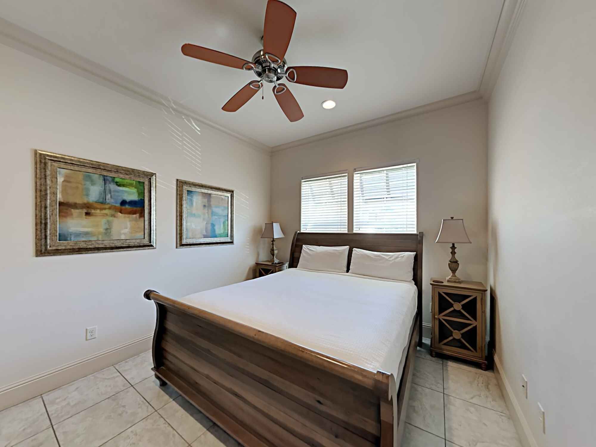 A bedroom with a wooden bed, two bedside tables with lamps, a ceiling fan, and two windows with blinds.