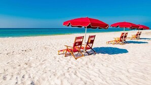 Une plage de sable blanc bordée d’une eau d’un bleu limpide. Des parasols rouges et des chaises de plage en bois sont disposés en rangées.