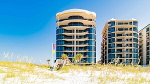 Un hôtel situé en bord de mer, doté de nombreux balcons, d’une plage de sable en contrebas et offrant une vue sur un ciel bleu dégagé.