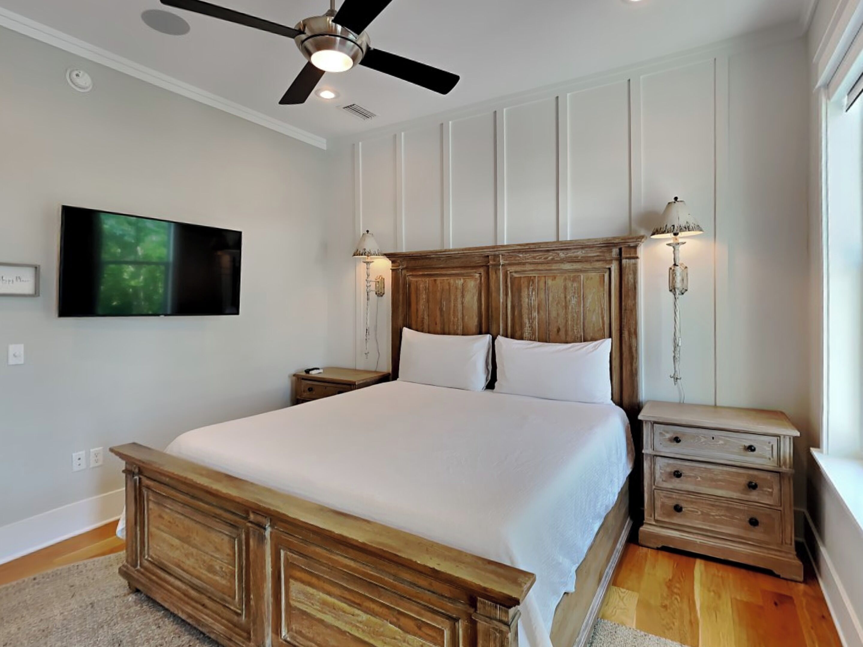 A bedroom with a wooden bed, a ceiling fan, bedside lamps, a TV, and a nightstand with drawers.