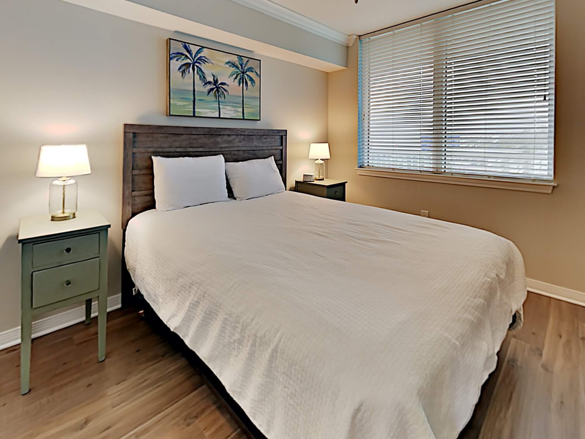 A bedroom with a large bed, two bedside tables with lamps, a wooden headboard, a painting of palm trees, and a window with blinds.