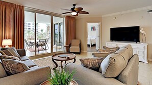 A spacious living room with a large sofa, a coffee table, and a television. There is a balcony with a view of palm trees and buildings.