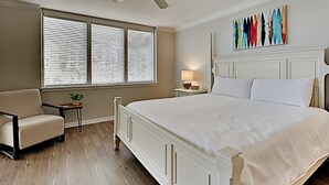 A bedroom with a large bed, a chair, a nightstand, a lamp, and a window with blinds.