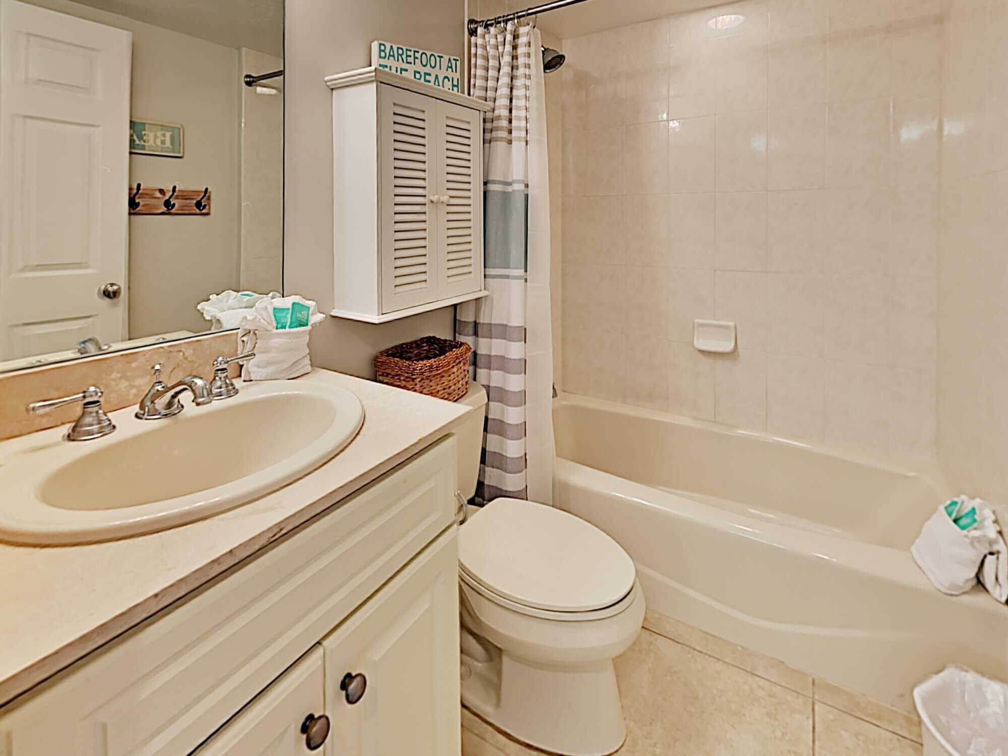 A bathroom with a sink, toilet, and bathtub. There is a sign above the bathtub that reads "Barefoot at the Beach."