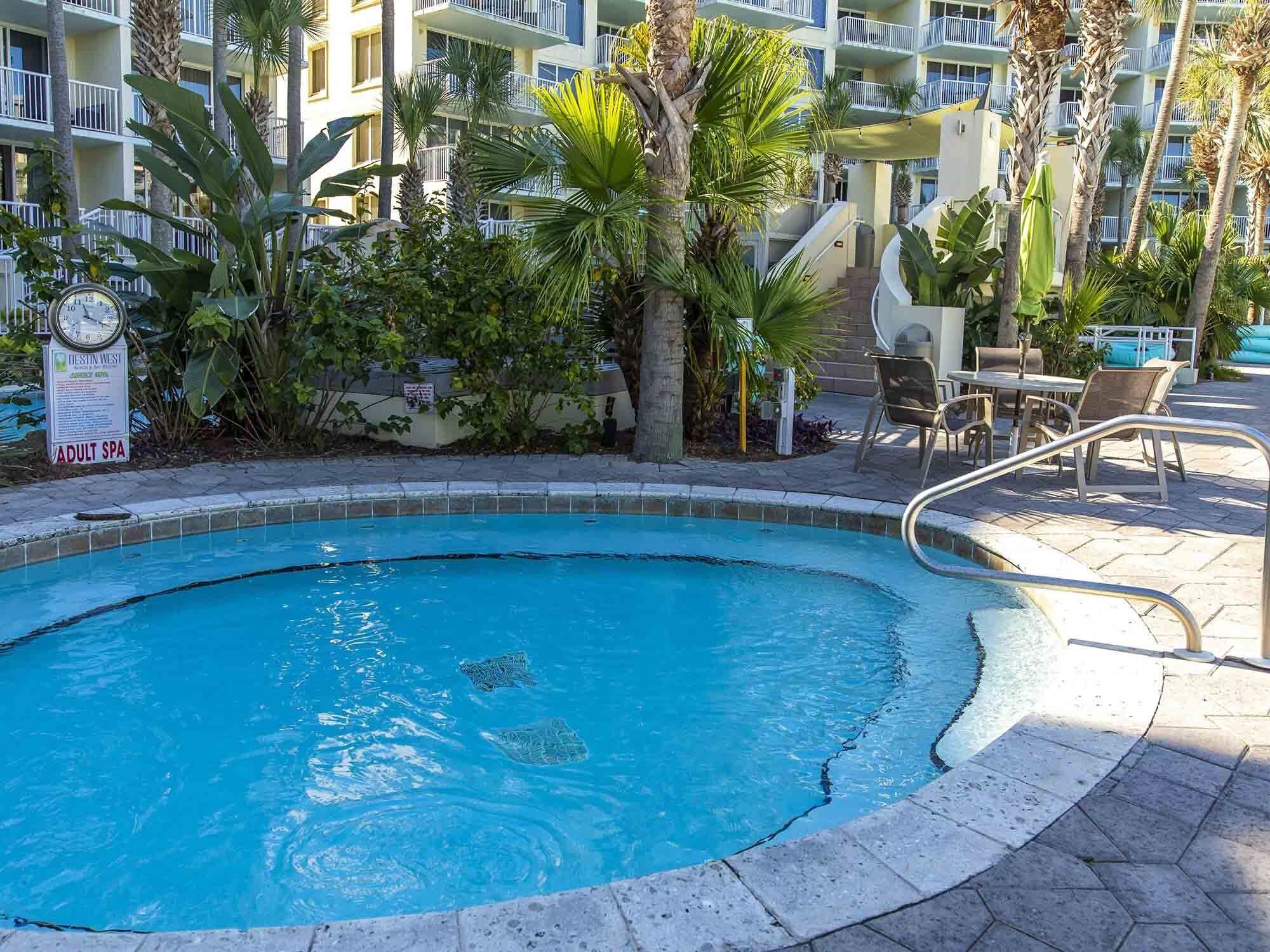 Una piscina con un reloj y señalización de spa, rodeada de palmeras y un edificio de varios pisos con balcones.