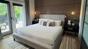 Un dormitorio moderno con una cama grande, un cabecero de madera con listones, dos mesitas de noche con lámparas y vistas a un espacio exterior a través de una puerta corrediza de vidrio.