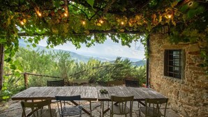 Een rustieke eethoek buiten, met een stenen muur, een houten tafel en metalen stoelen, met uitzicht op een bergachtig landschap.