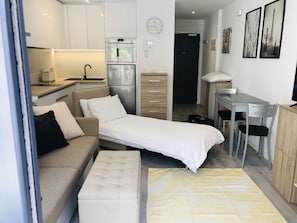 Quarto compacto e moderno com cama, sofá, pequeno espaço de cozinha e escrivaninha.