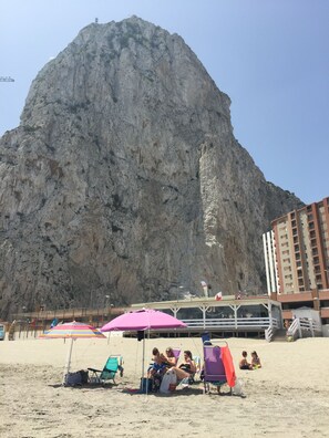A beach with a large rock formation in the background, beach chairs, umbrellas, and a building with a balcony.