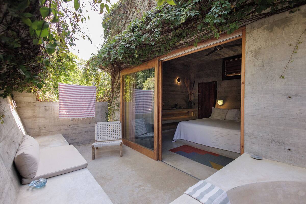 A cozy outdoor bedroom with a glass door leading to a bed, a small seating area, and a view of the surrounding greenery.