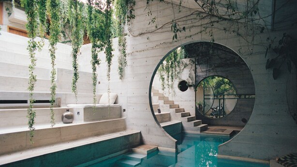 Uma área interna de piscina moderna com uma janela circular de observação, degraus de pedra e plantas pendentes.