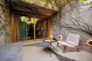 Un rincón exterior acogedor con una silla de mimbre y una mesita, un vidrio que da a una habitación, y una pared de concreto con enredaderas.