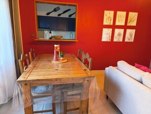 A dining area with a wooden table, chairs, a vase with flowers, and a kitchen in the background.