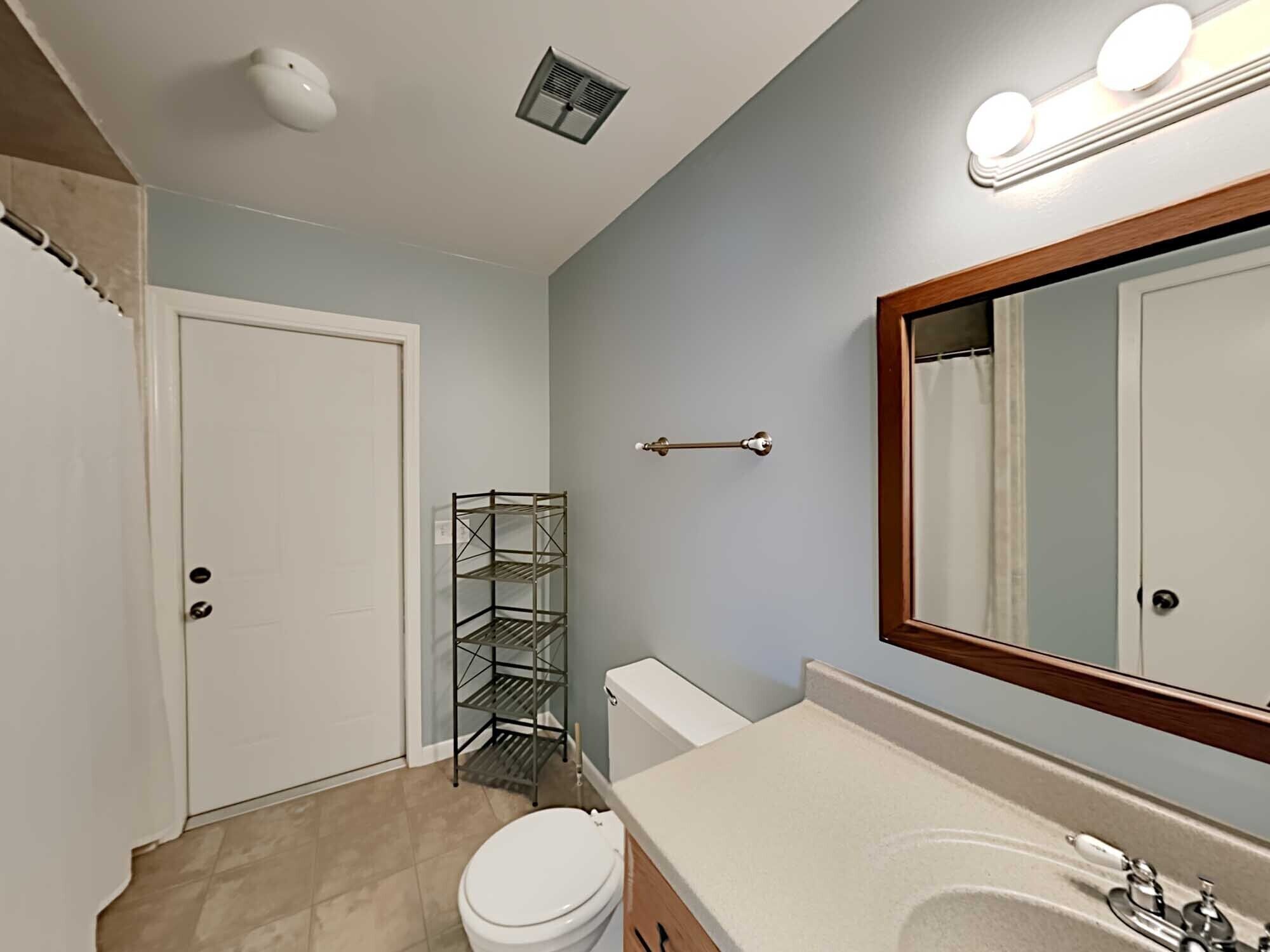 A bathroom with a toilet, sink, mirror, and a metal shelf.