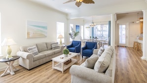 A spacious living room with a beige sofa, a blue armchair, and a coffee table. There is a dining area with a table and chairs, and a kitchen in the background. - Sandy Retreat by Southern Vacation Rentals (Destin)