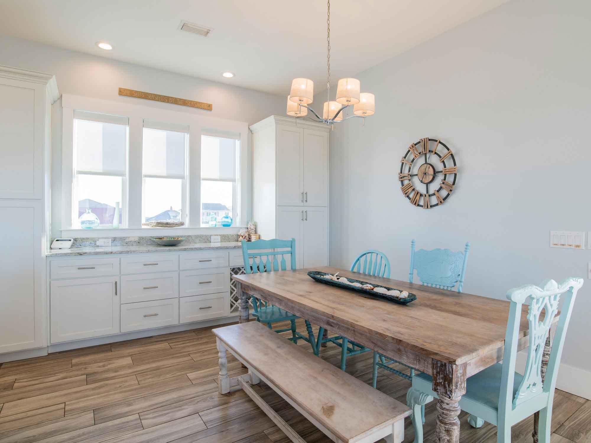 Una cocina con armarios blancos, una mesa de comedor de madera, sillas color turquesa y un reloj de pared.