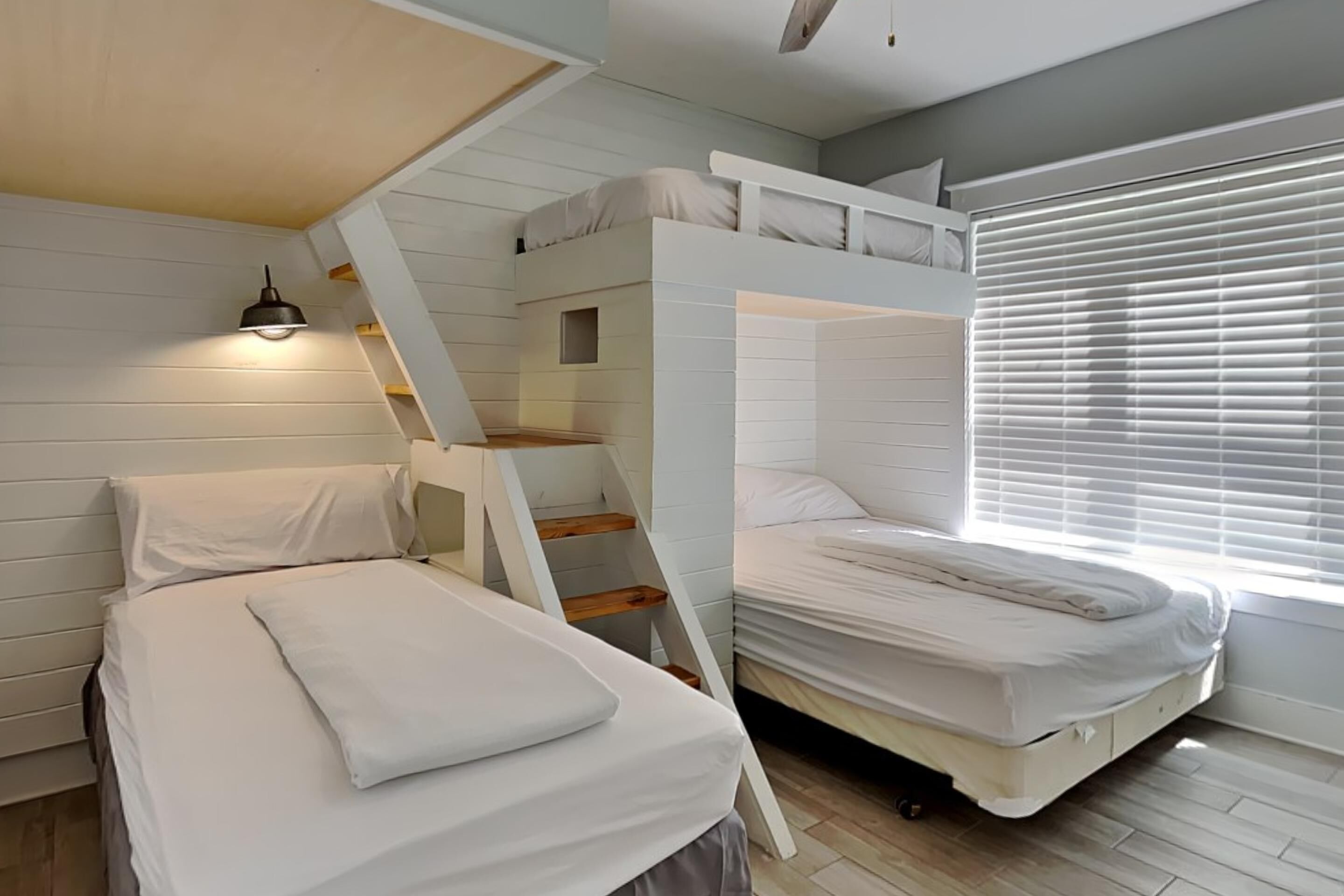 A room with bunk beds, a ladder, a bedside table, and a window with blinds.