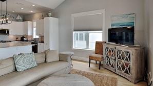 A modern kitchen with white cabinets, a dining area with a round table, a sofa with patterned cushions, and a television on a wooden cabinet. - Magnolia House by Southern Vacation Rentals (Santa Rosa Beach)