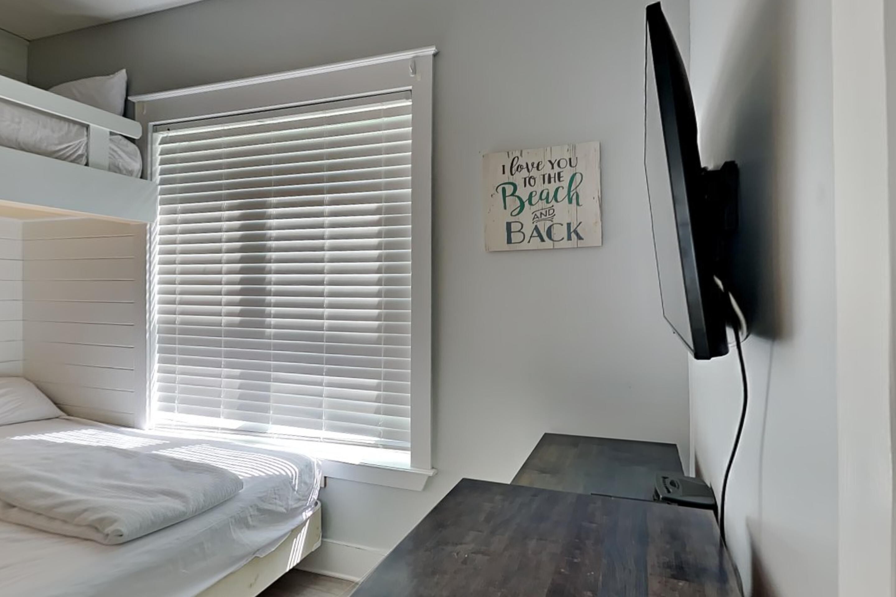 A bedroom with a bunk bed, a large window with blinds, a flat-screen TV mounted on the wall, and a small wooden table.