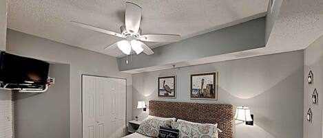 A hotel room with a bed, a ceiling fan, a TV mounted on the wall, a door, and two bedside tables with lamps.