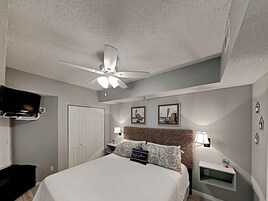 Habitación de hotel con cama, ventilador de techo, televisor montado en la pared, puerta y dos mesitas de noche con lámparas.