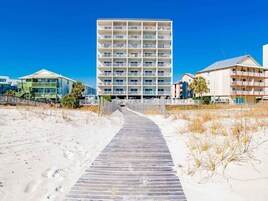 Un hotel frente al mar con un edificio central que cuenta con varios balcones, un paseo marítimo que conduce a la entrada y una zona de playa arenosa.