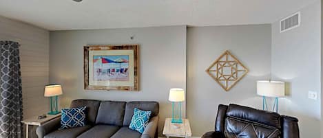 A living room with a gray sofa, a black leather chair, two side tables with lamps, a framed beach picture, and a geometric wall decoration.
