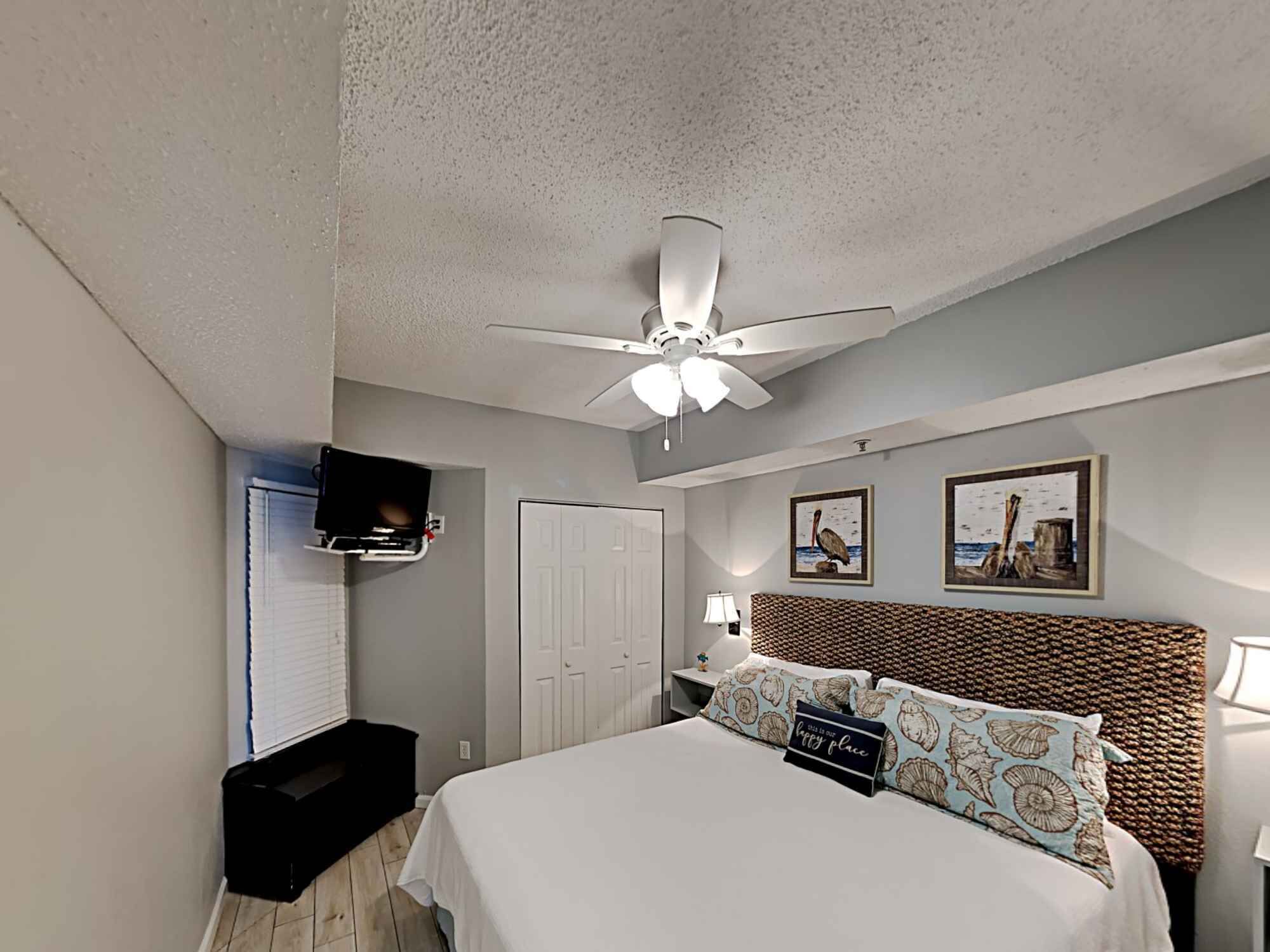 A hotel room with a bed, a ceiling fan, a television mounted on the wall, and two framed pictures on the wall.