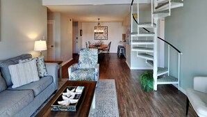 A spacious living room with a grey sofa, a coffee table with decorative items, a dining area with a chandelier, and a white spiral staircase. - Cotton Bayou Condominiums by Southern Vacation Rentals (Orange Beach)
