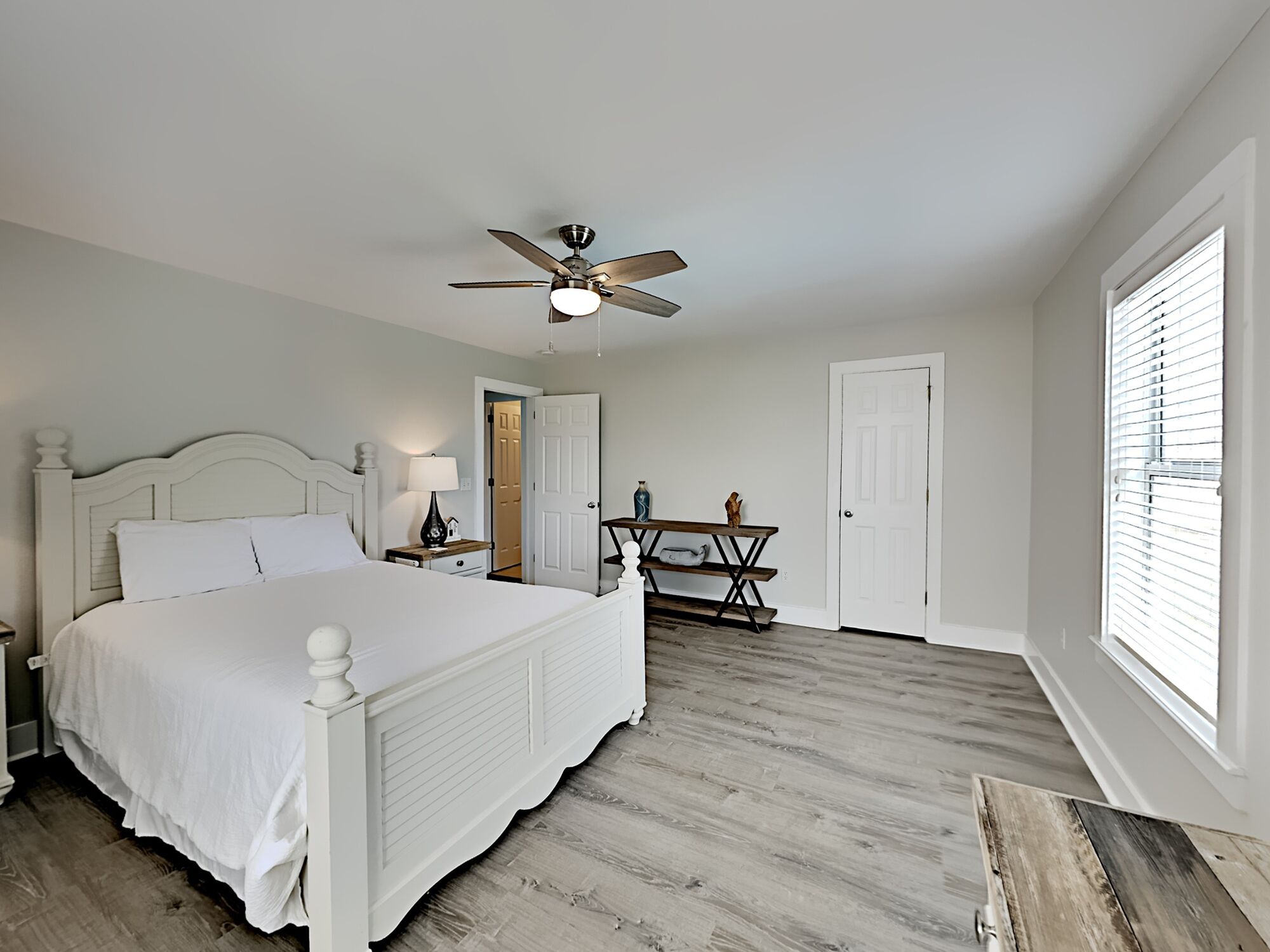 A bedroom with a white bed, a ceiling fan, a wooden dresser, and a door leading to another room.