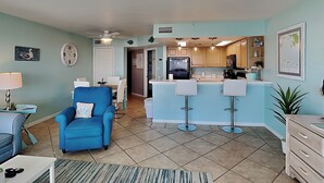Ein geräumiges Wohnzimmer mit blauem Sessel, einem Essbereich mit Tisch und Stühlen, einer Küche mit Bar und einem Deckenventilator.