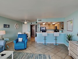 A spacious living room with a blue armchair, a dining area with a table and chairs, a kitchen with a bar area, and a ceiling fan.