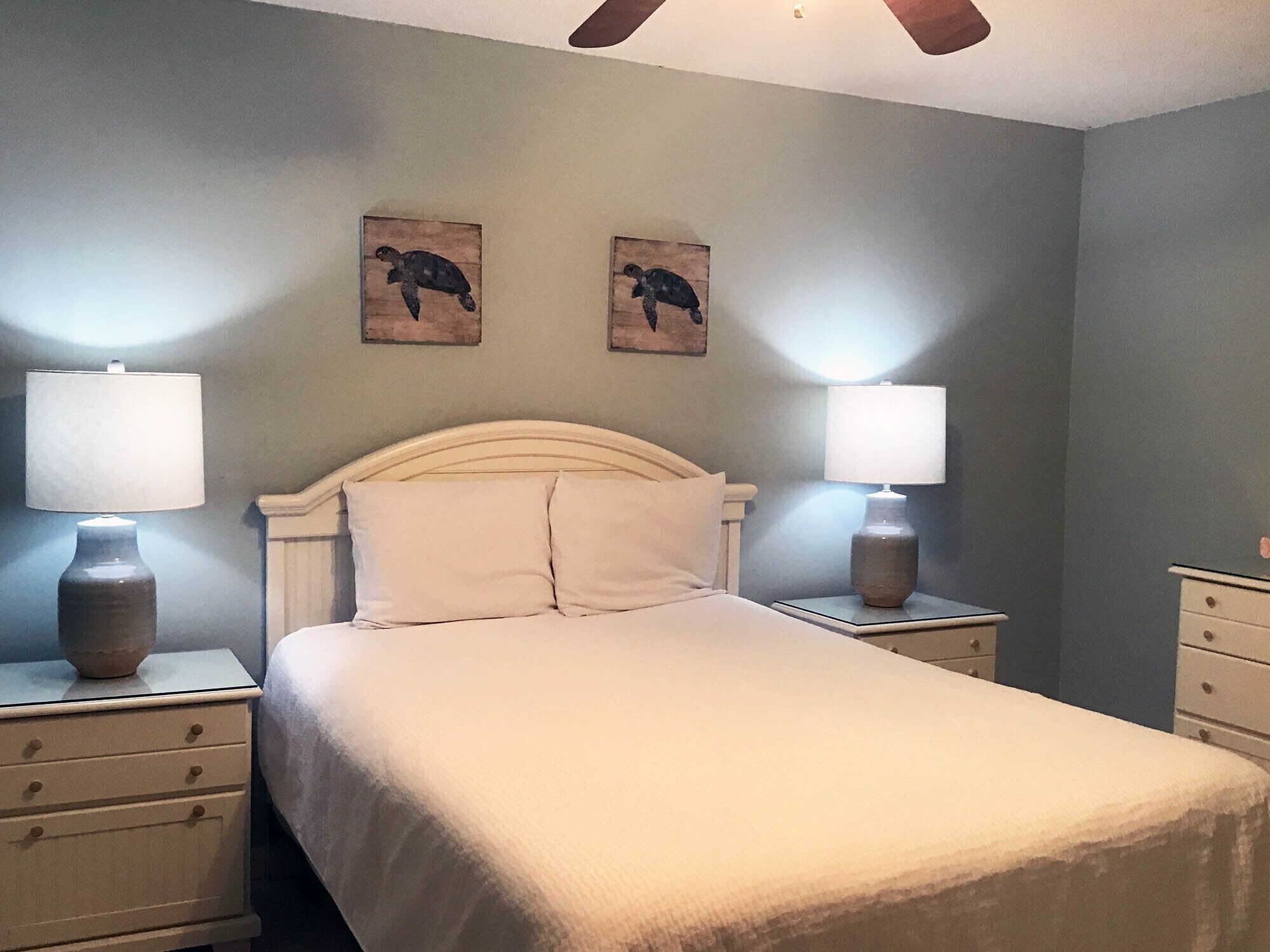 Quarto com uma cama, duas mesas de cabeceira, duas luminárias e duas obras de arte de tartarugas nas paredes.
