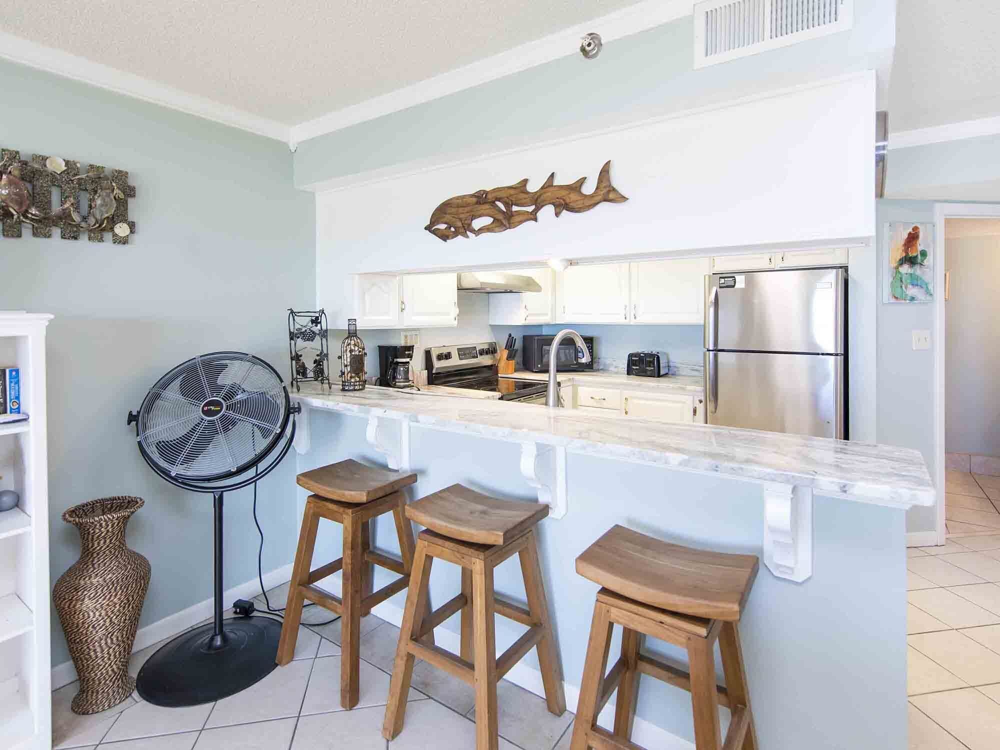 A modern kitchen with a marble countertop, wooden stools, a fan, and a refrigerator.