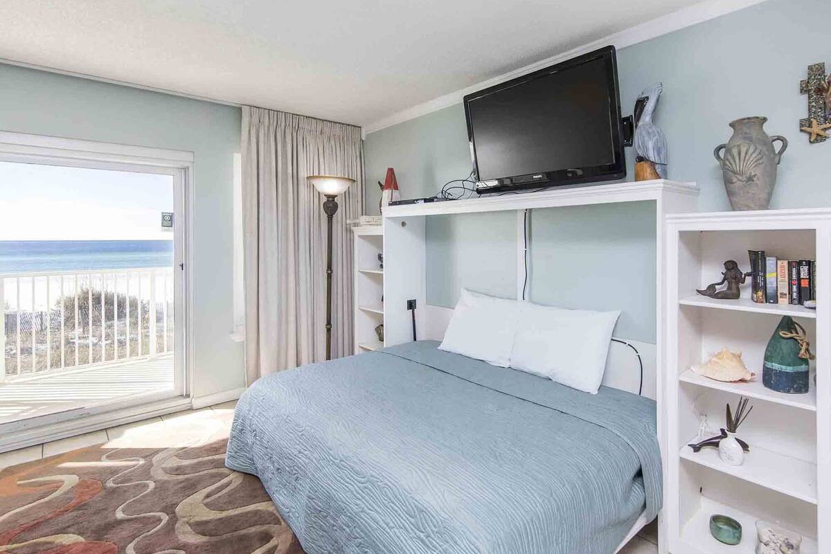 A bedroom with a bed, a television, a bookshelf, and a view of the ocean.