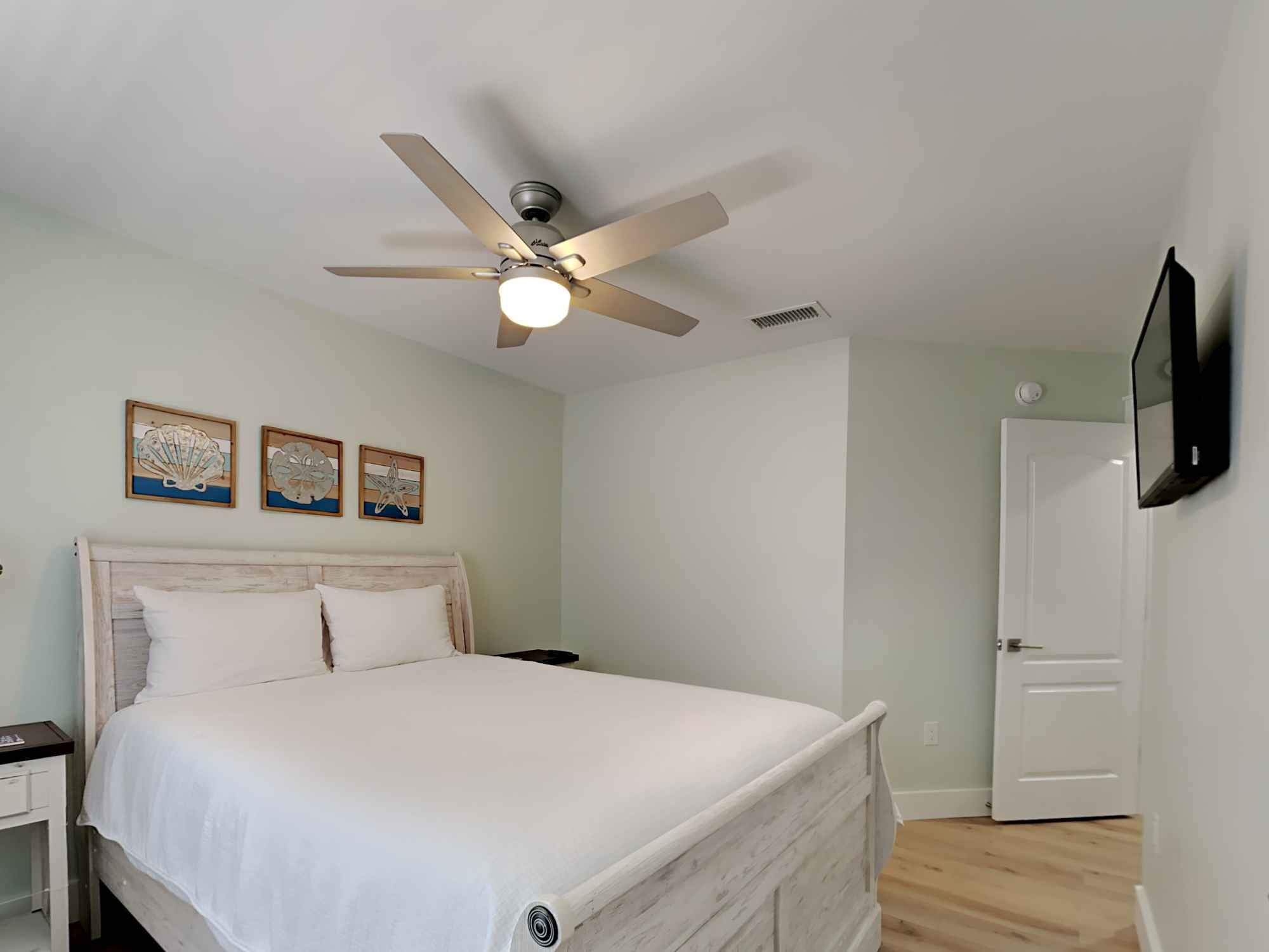 Une chambre à coucher avec un lit, un ventilateur de plafond, un téléviseur fixé au mur et trois tableaux encadrés accrochés au mur.
