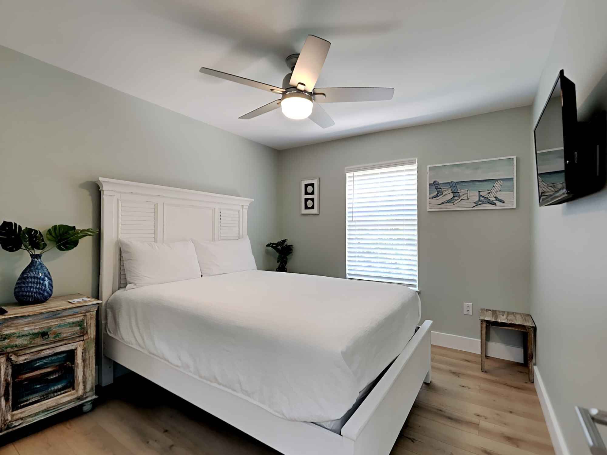 A bedroom with a white bed, a ceiling fan, a window with blinds, and a small table with a plant.