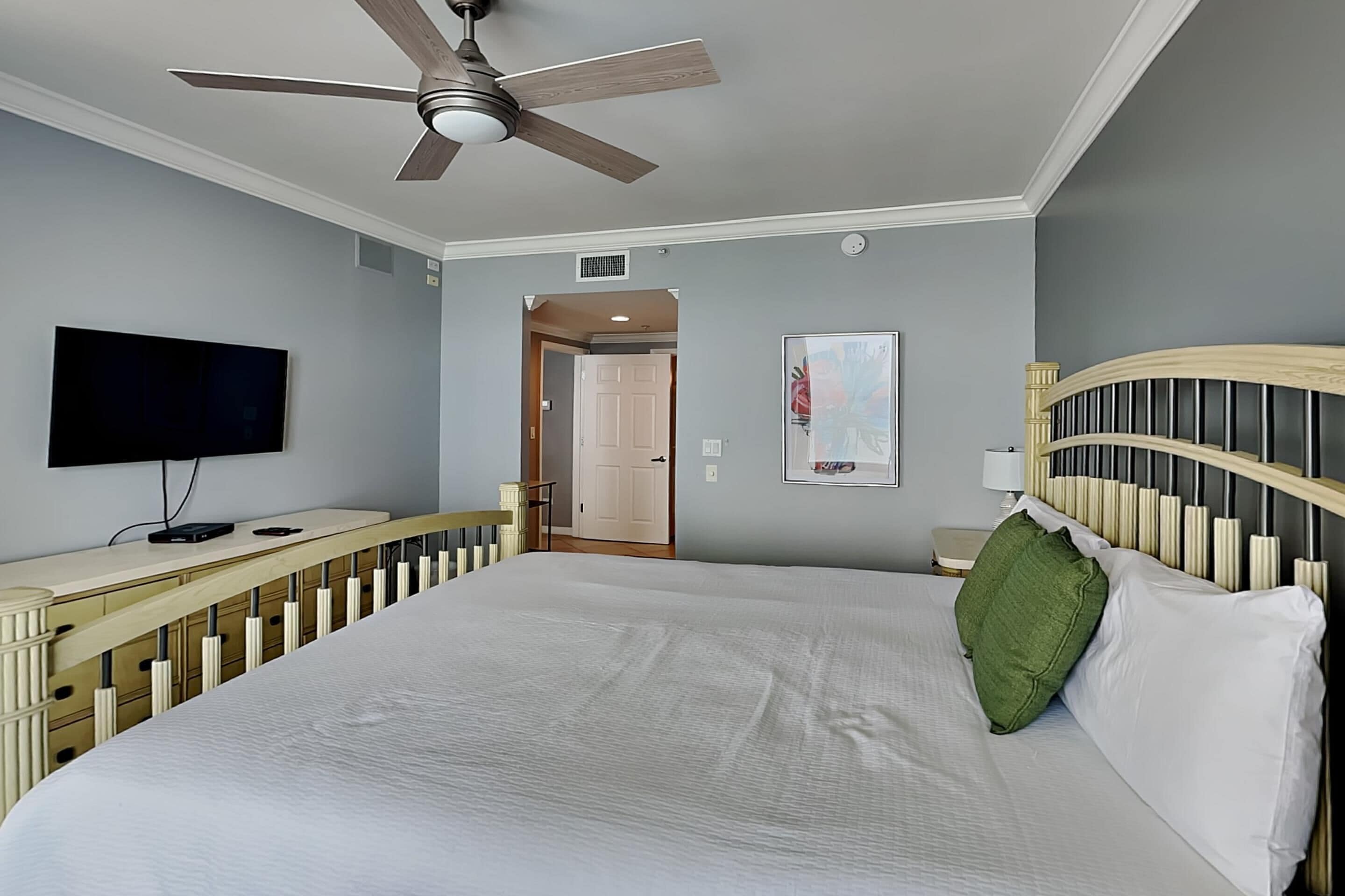 A hotel room with a large bed, a ceiling fan, a flat-screen TV, a wooden headboard, and a painting on the wall.