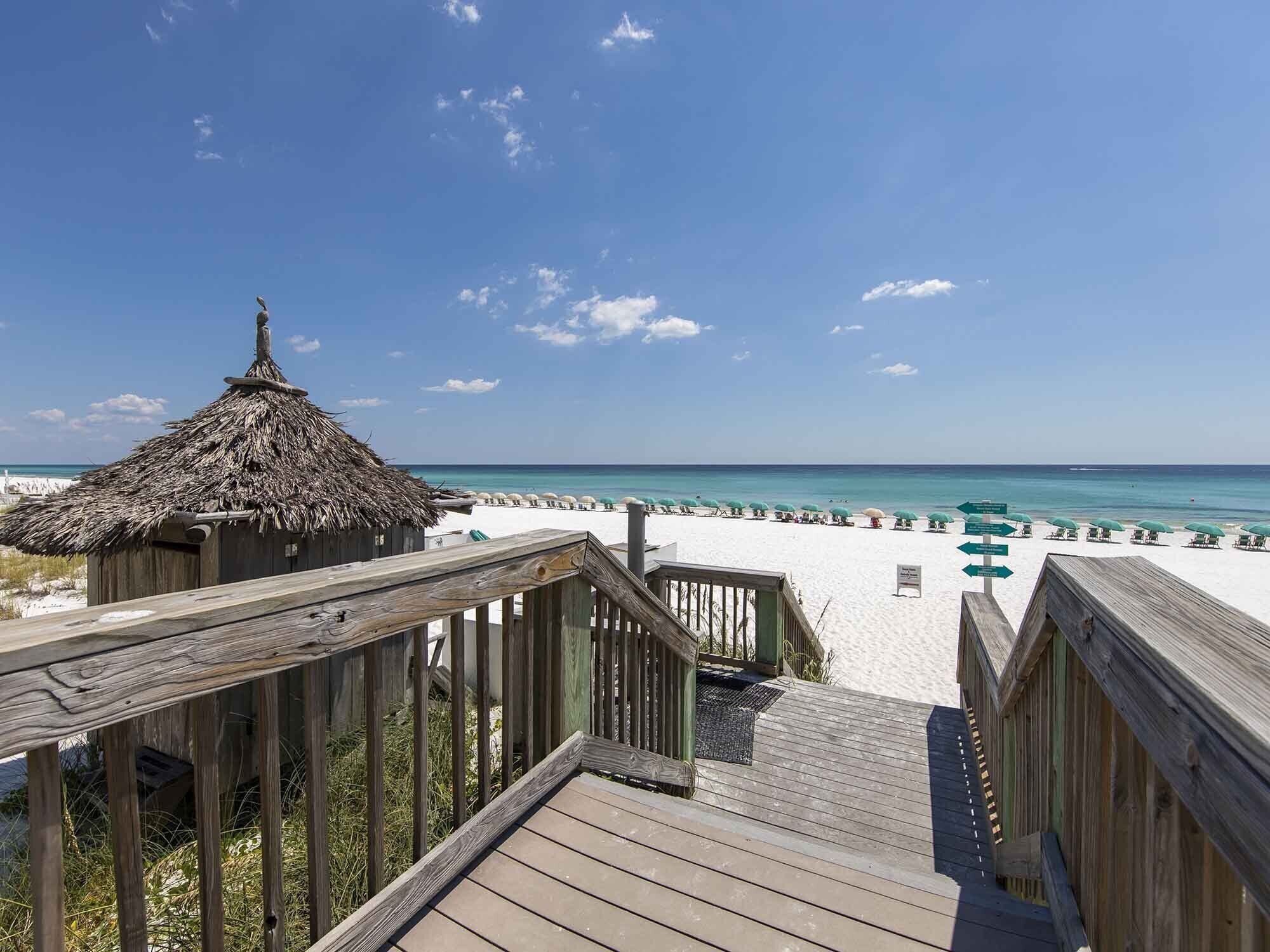 A wooden deck leading to a beach with a thatched-roof structure, white sand, and turquoise water.
