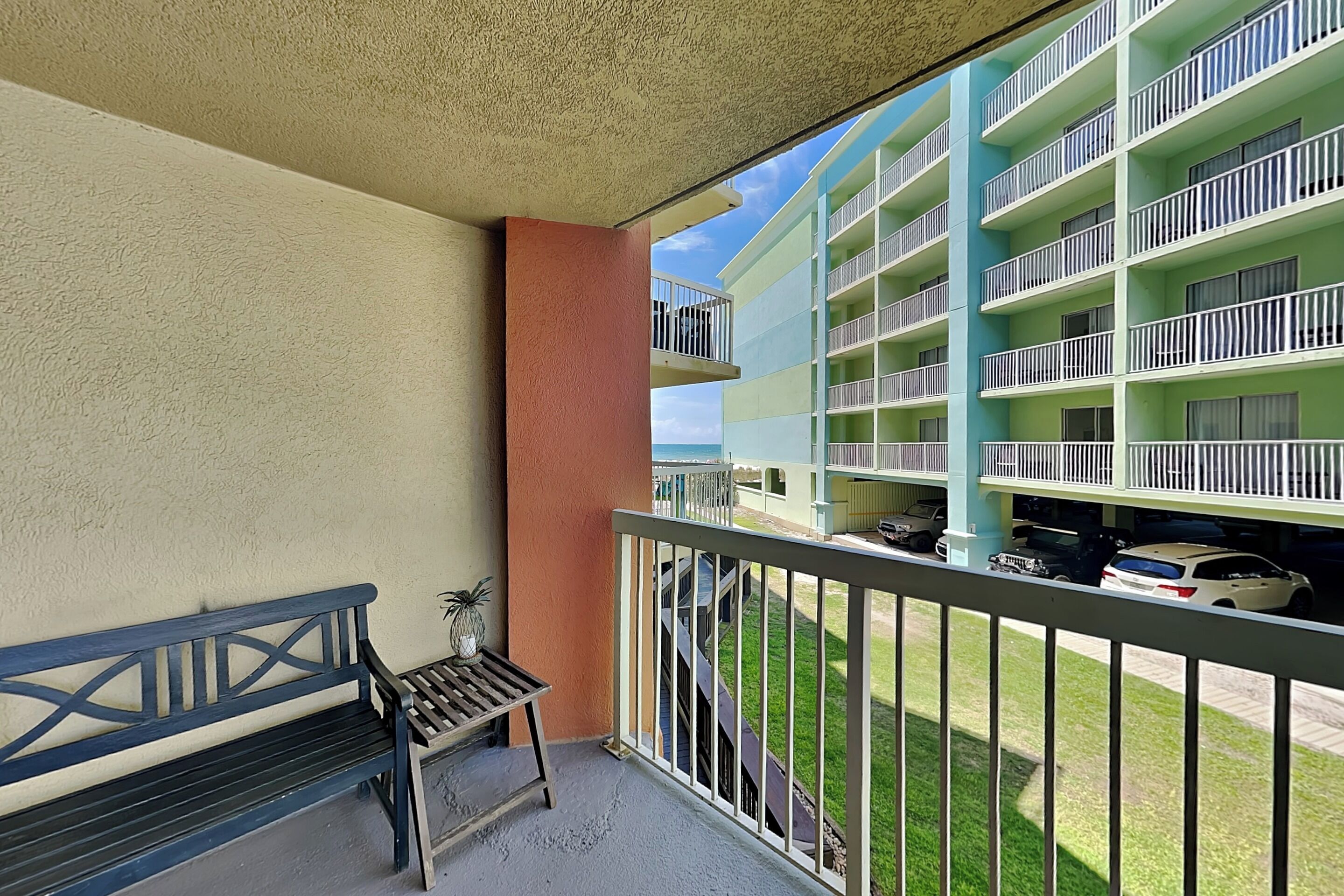 Een balkon met een bankje en een kleine tafel, met uitzicht op een gebouw met meerdere balkons en geparkeerde auto's.