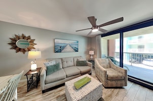 A living room with a sofa, armchair, and a decorative coffee table. There's a ceiling fan, a wall clock, and a painting of a pier.