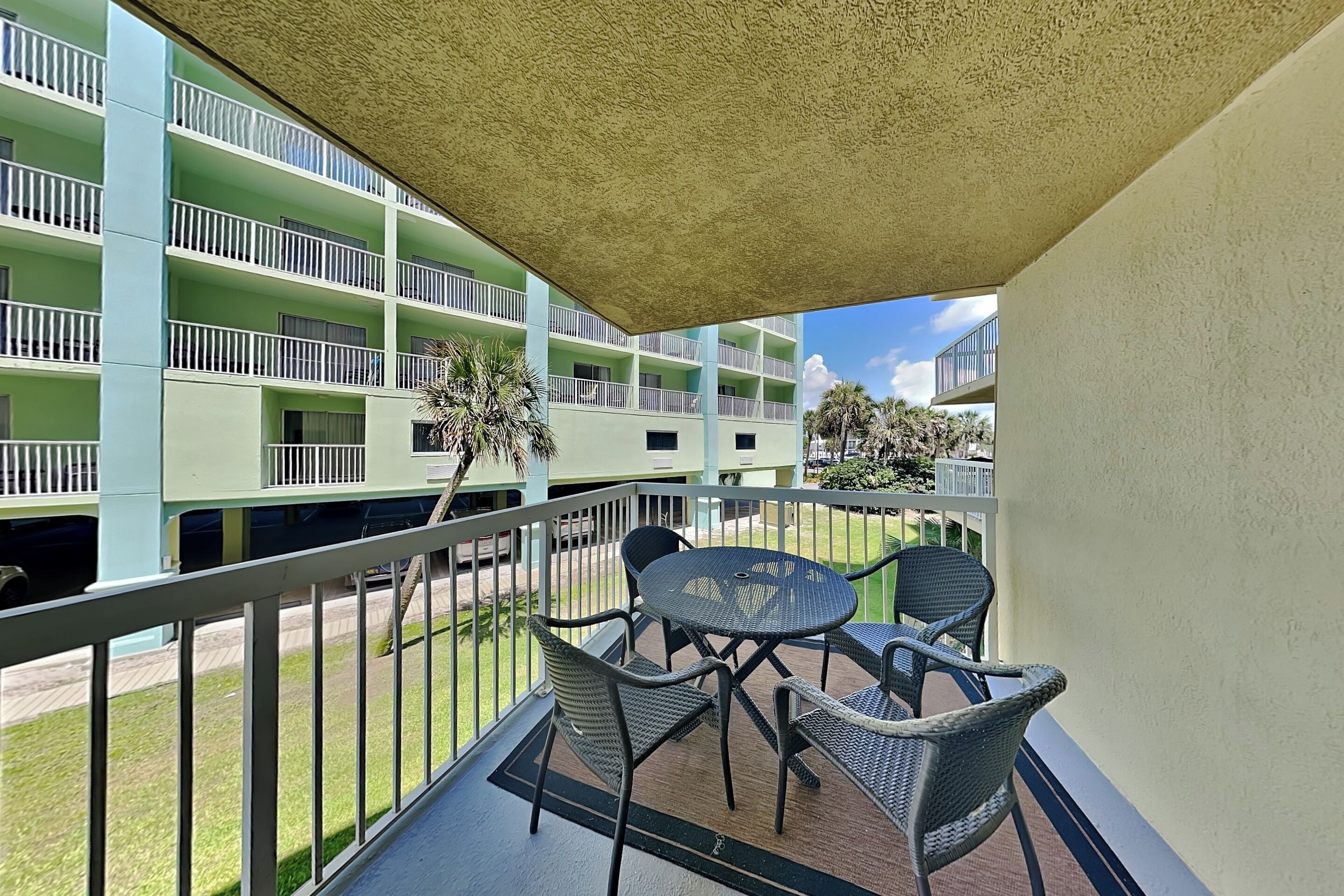 A balcony with a table and chairs, a view of a multi-story building with balconies, and a clear sky.