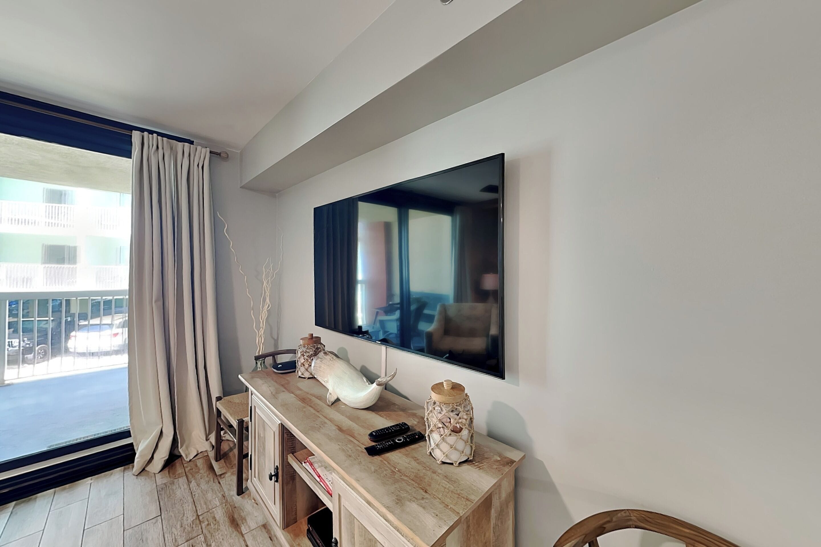 Ein modernes Wohnzimmer mit einem großen Flachbildfernseher an der Wand, einem Holzkonsolentisch mit Dekoration, einem Sessel und Blick auf die Straße durch ein großes, mit Vorhängen drapiertes Fenster.