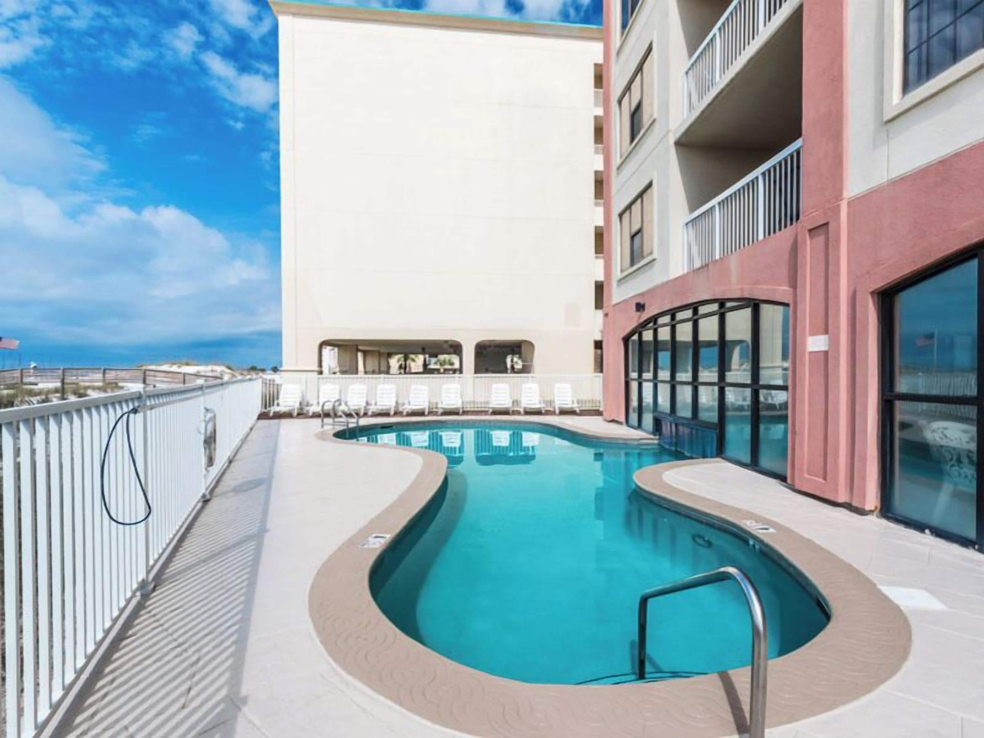 A rooftop pool with a clear view of the ocean, surrounded by a white railing and a building with balconies.