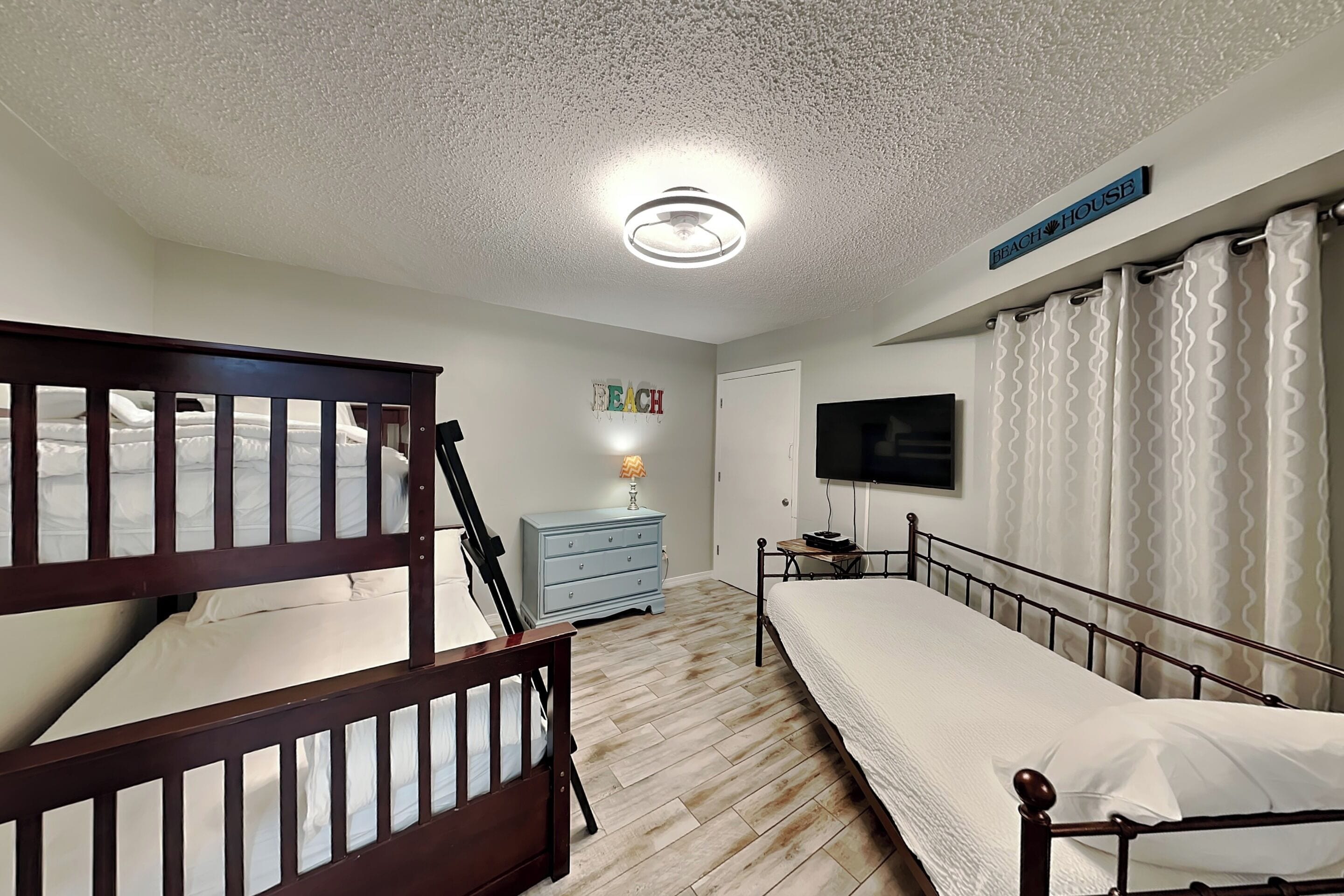 A bedroom with bunk beds, a dresser, a television, and a sign that says 'BEACH HOUSE'.