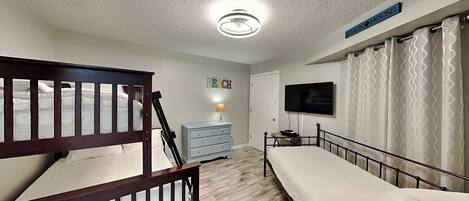 A bedroom with bunk beds, a dresser, a television, and a sign that says 'BEACH HOUSE'.