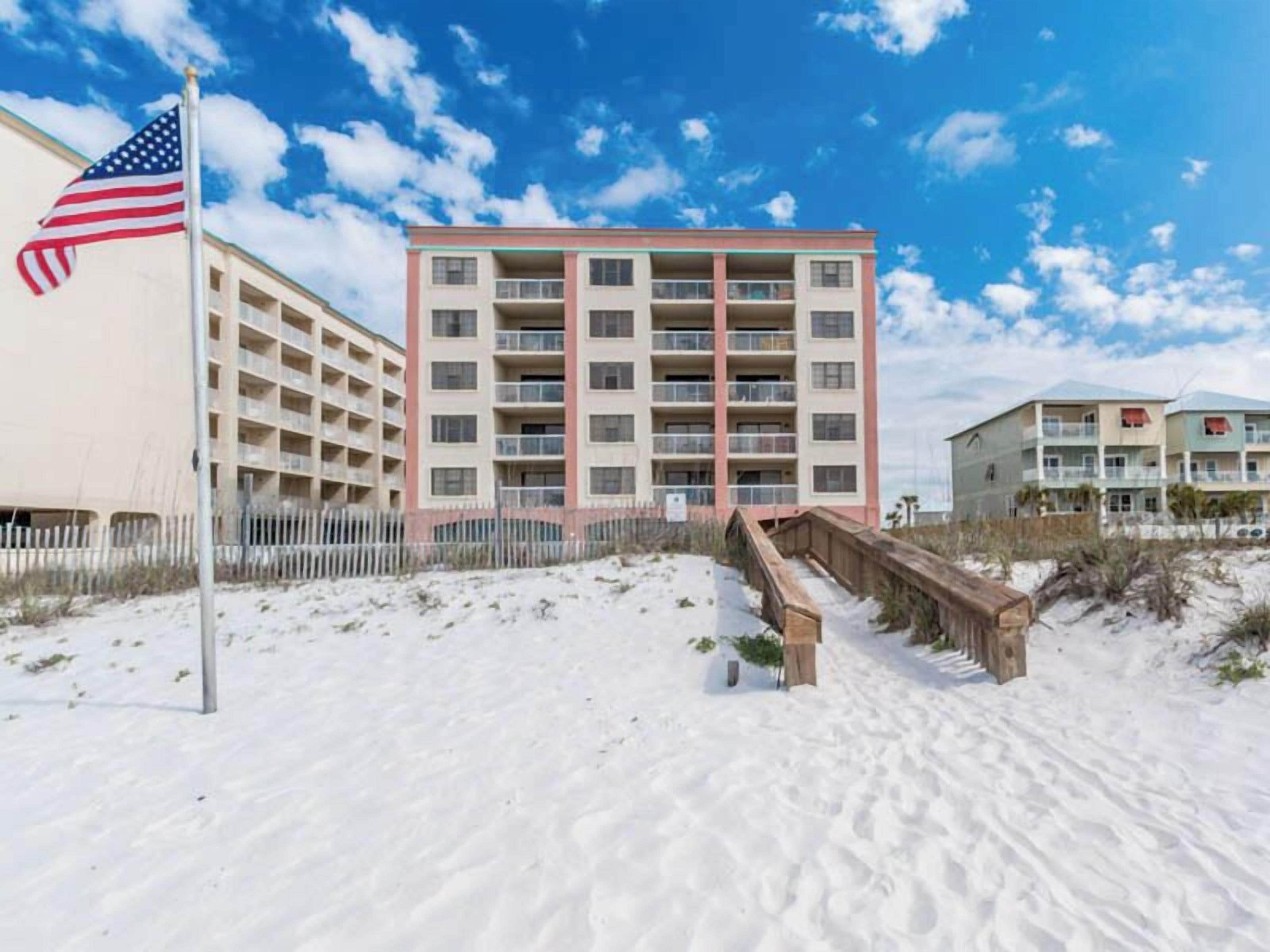 A multi-story hotel with a central courtyard, a flagpole with an American flag, and a sandy beach area.